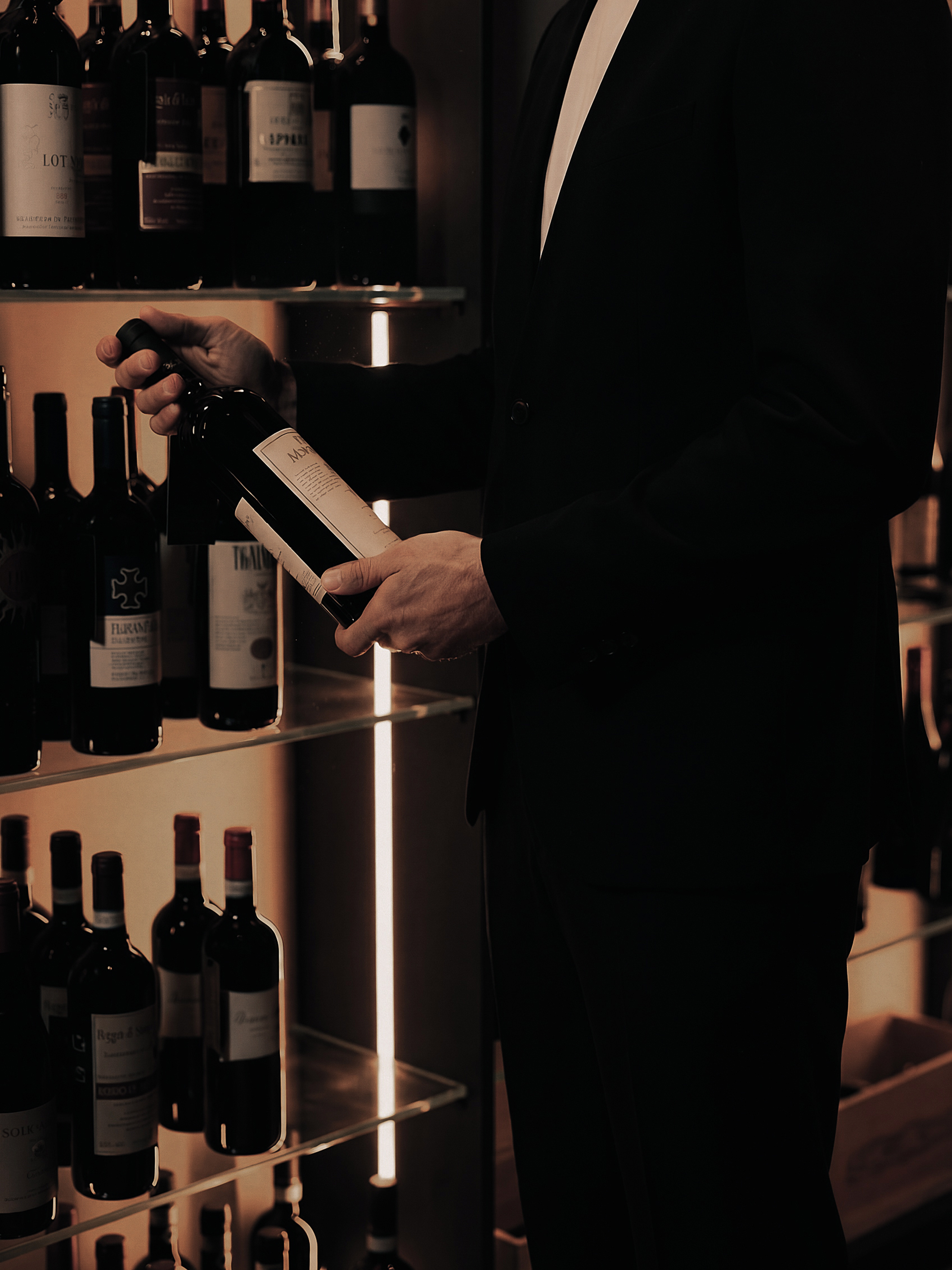 Mani che presentano una bottiglia di vino con etichetta in primo piano, sullo sfondo scaffali illuminati della cantina QOR